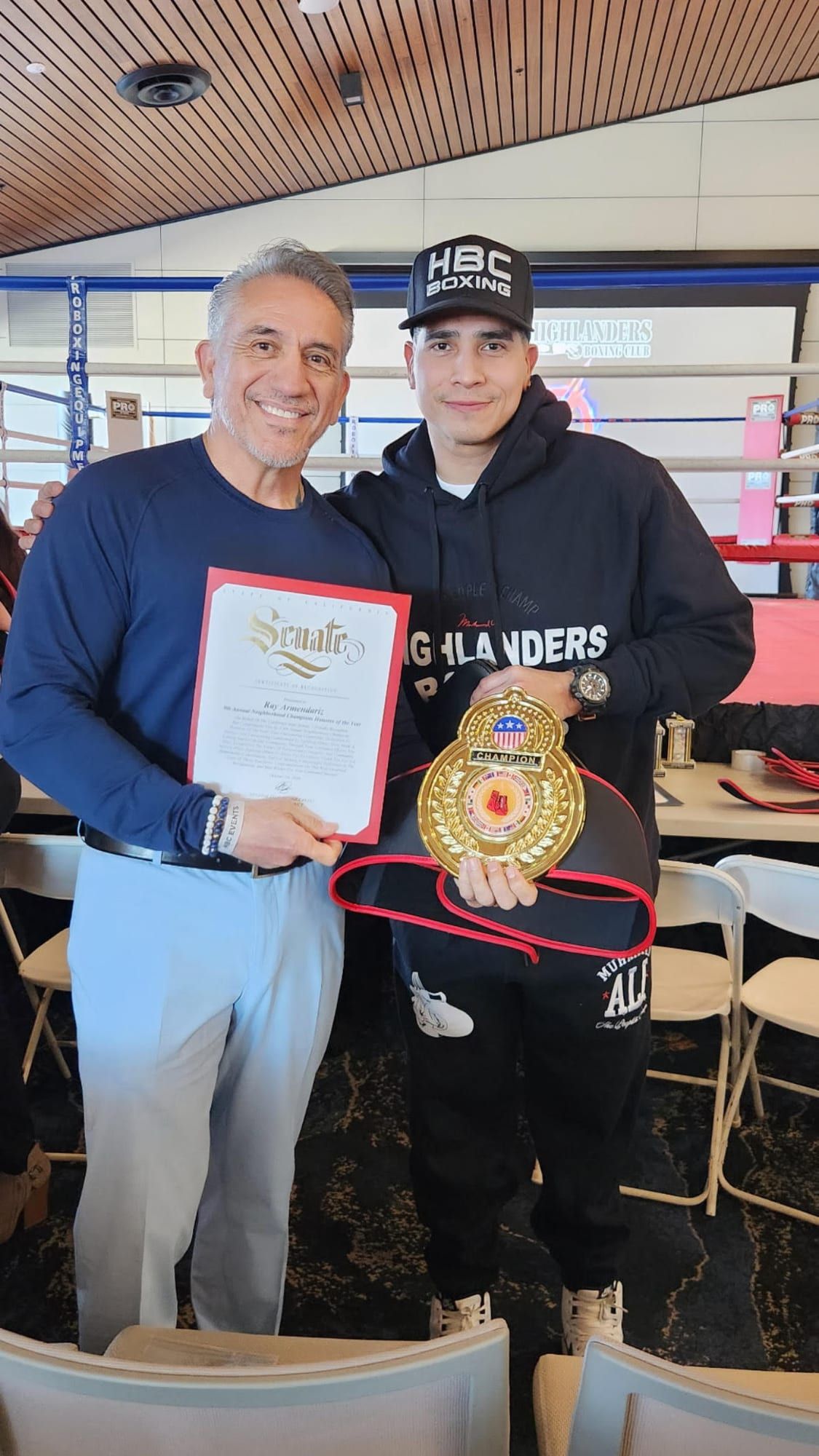 Two men in front of the ring, one holding a California State Senate certificate of recognition and the other a champion belt