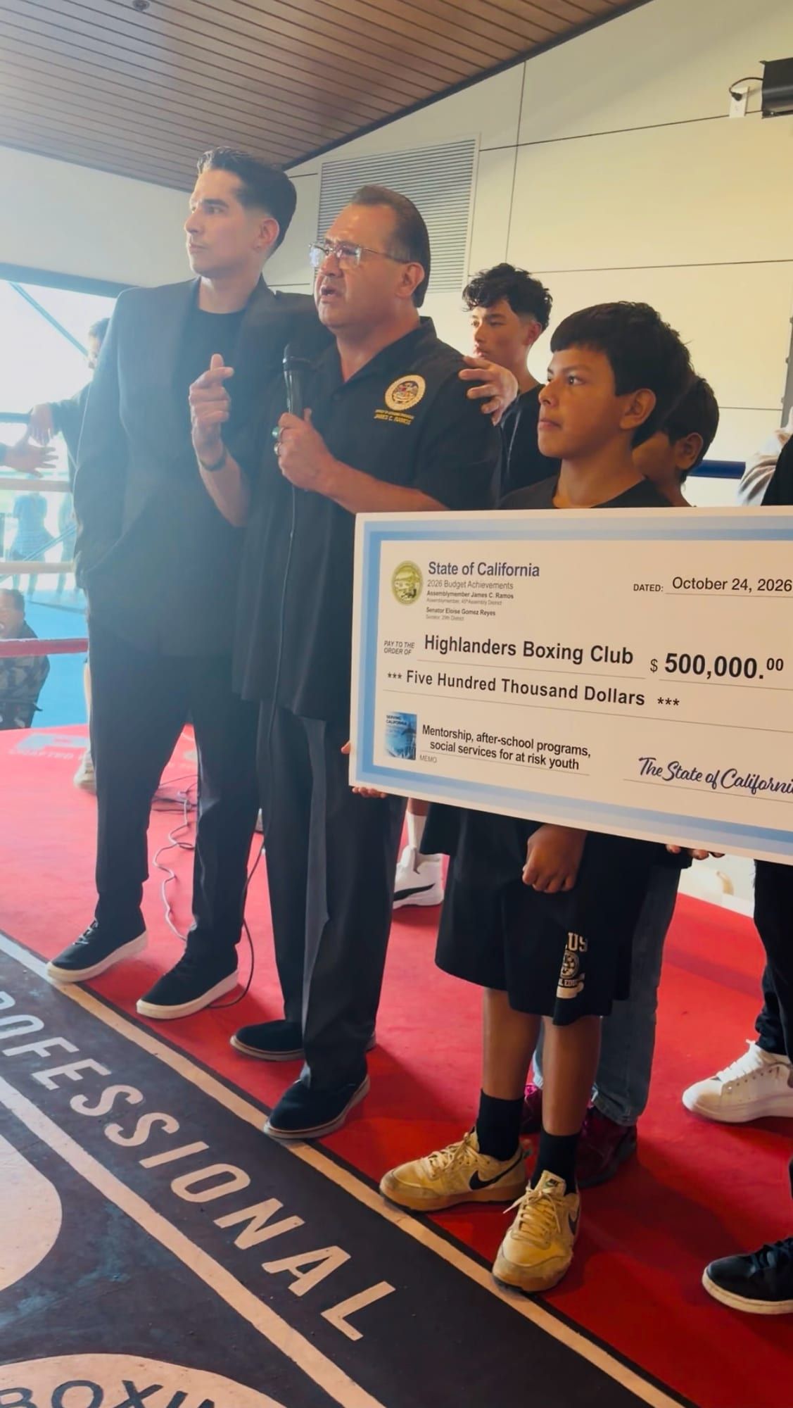 A speaker with a microphone in the ring, beside youth holding the $500,000 State of California check